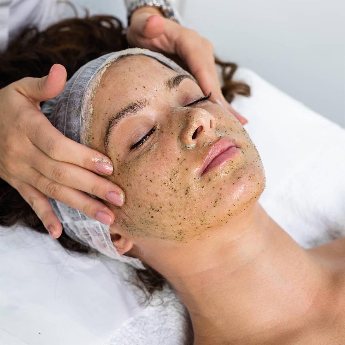 Person receiving a decaar algae peel facial treatment with a green, textured mask applied to their face, while lying down and having their head gently massaged.