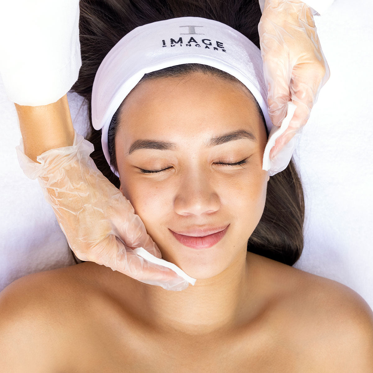 Person receiving a facial treatment with gloved hands applying cotton pads to their cheeks.