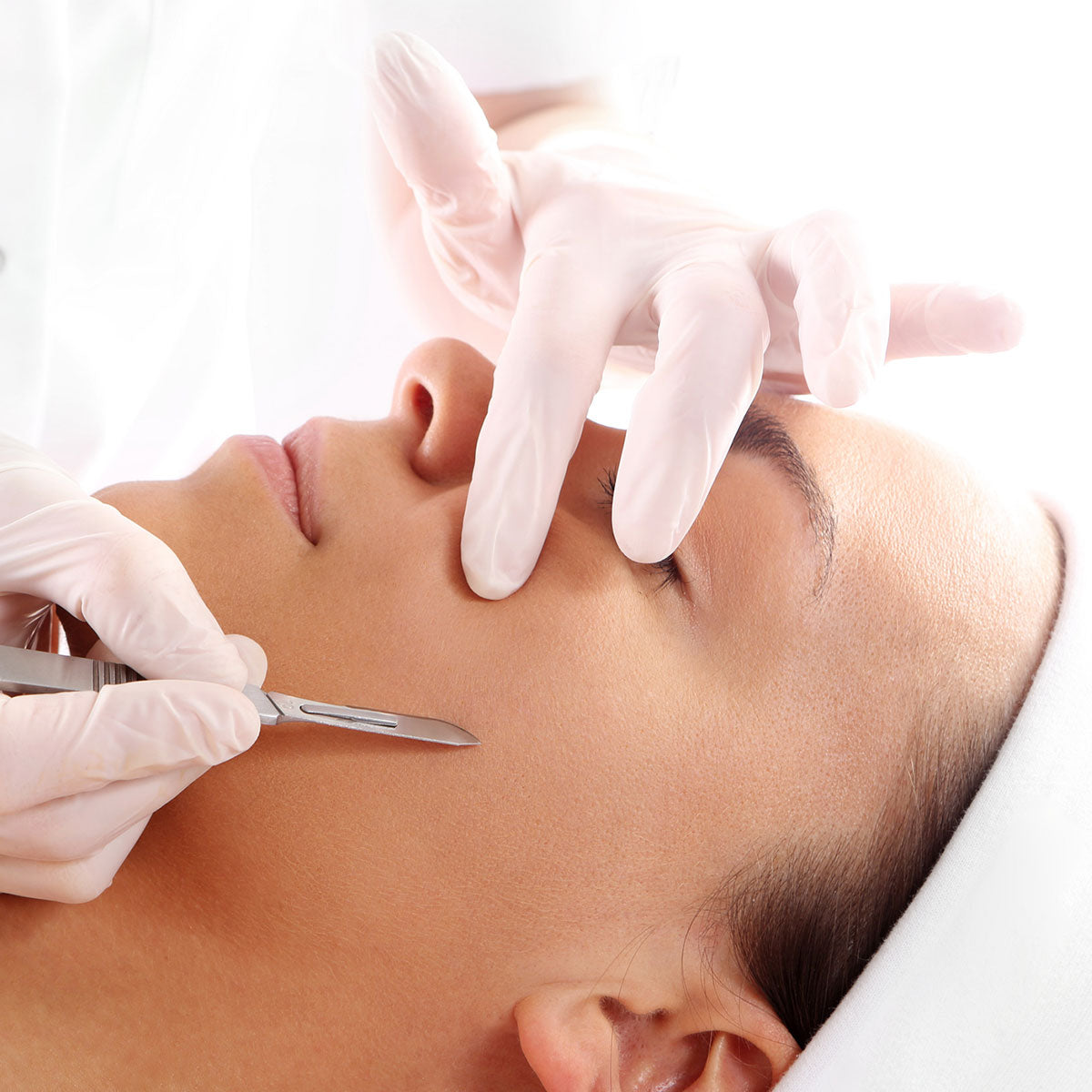 Person receiving a facial treatment with gloved hands using a scalpel on their cheek.